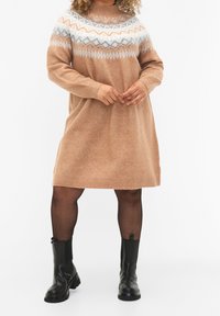 Robe pull en marron clair avec un empiècement décoré de blanc et de gris. Manches longues, coupe décontractée et poignets côtelés. Portée avec des bottes noires.