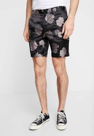 Mand iført sorte shorts med blomsterprint, hvid t-shirt og sorte lave sneakers med hvide snørebånd, stående mod en ensfarvet baggrund.