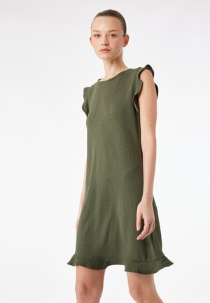 Donna con capelli raccolti che indossa un abito verde oliva al ginocchio con maniche e orlo arricciati, su sfondo bianco semplice.