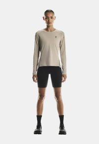 Beige Sportoberteil mit langen Ärmeln, Rundhalsausschnitt und Logo, kombiniert mit schwarzen Shorts bis zur Mitte des Oberschenkels, getragen mit schwarzen Socken und Laufschuhen.