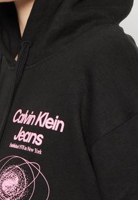 Svart hoodtröja i bomull med en rosa logotyp "Calvin Klein Jeans" och grafiska detaljer. Inkluderar en huva med dragsnodd och synlig textur.