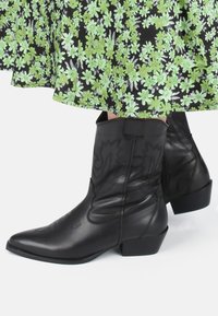 Botas de vaquero de cuero negro con punteras puntiagudas, tacones cuadrados y costuras decorativas. Suave textura con cierre de cremallera en el lateral.