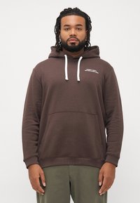 Hombre con barba y rastas, vistiendo una sudadera marrón con cordones blancos y pantalones verdes oliva, de pie frente a un fondo sencillo.
