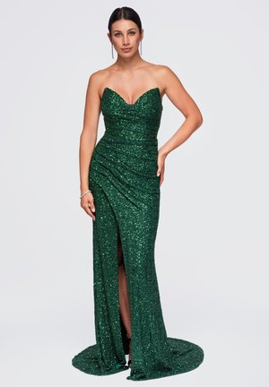 Femme portant une robe bustier vert foncé scintillante avec une fente haute sur la cuisse, cheveux foncés attachés, posant avec une main sur la hanche et l'autre sur le côté.