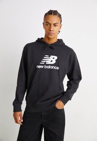 Fekete kapucnis pulóver kenguru zsebbel és húzózsinórral. Elöl található egy nagy fehér "New Balance" logó. Sima szövettextúra.