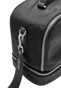 Róg czarnej torby materiałowej z srebrnym suwakiem i metalowym klamrą przymocowanym do czarnego regulowanego paska.