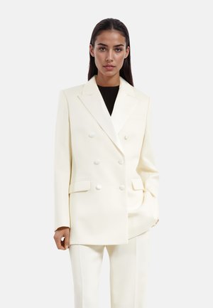 Blazer de doble botonadura en color crema con solapas de pico, textura suave y botones en contraste. Silueta ajustada con bolsillos laterales funcionales.