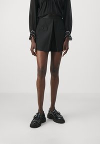 Shorts noirs au design ajusté avec plis, deux poches avant avec boutons, associés à des mocassins noirs vernis. Texture de tissu lisse.