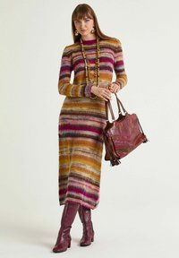 Gestreiftes Strickkleid in warmen Gelb-, Pink- und Lilatönen, kombiniert mit einer burgunderfarbenen, strukturierten Handtasche und kniehohen burgunderfarbenen Stiefeln.