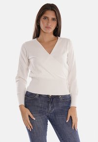 Maglione avvolgente bianco con maniche lunghe e orlo a coste, realizzato in materiale morbido, abbinato a jeans blu scuro. Il modello posa su uno sfondo neutro.