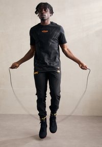 Camiseta atlética negra con logo naranja de Venum, combinada con pantalones negros que presentan el logo amarillo de UFC, y zapatillas altas negras texturizadas.
