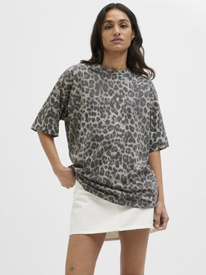 Naine, kes kannab halli leopardimustriga üle mõõdu suure T-särgi ja valget miniseelikut, seisab ühe käe taskus ja neutraalse väljendiga.