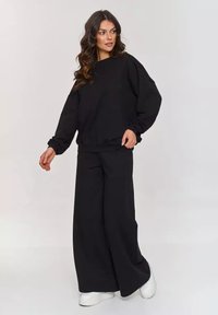 Czarna, oversize'owa bluza z balonowymi rękawami zestawiona z szerokimi czarnymi spodniami, wykonana z miękkiego materiału, noszona z białymi trampkami.