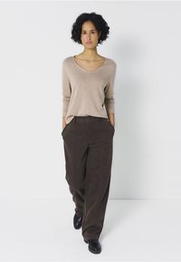 Pull beige à manches longues et col en V associé à un pantalon large marron. Le mannequin se tient en toute confiance avec les mains dans les poches, portant des chaussures noires.