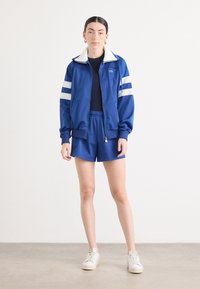 Jaqueta desportiva azul com fecho e riscas brancas, com mangas curtas, combinada com calções azuis a condizer, completada com sapatilhas brancas.