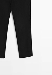 Zwarte broek met rechte pijpen en een kleine split aan de onderzoom op een witte achtergrond, alleen het onderste deel zichtbaar.