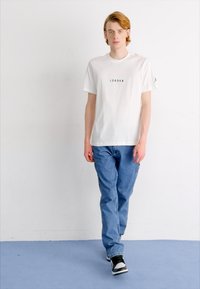 Camiseta de algodón blanca con un pequeño logo negro en el pecho, combinada con jeans de mezclilla azul y zapatillas negras y blancas. Fondo liso.