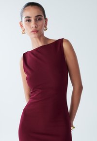 Burgundy sleeveless dress with a structured neckline, featuring a smooth texture and fitted silhouette. Gold statement earrings complement the look.
