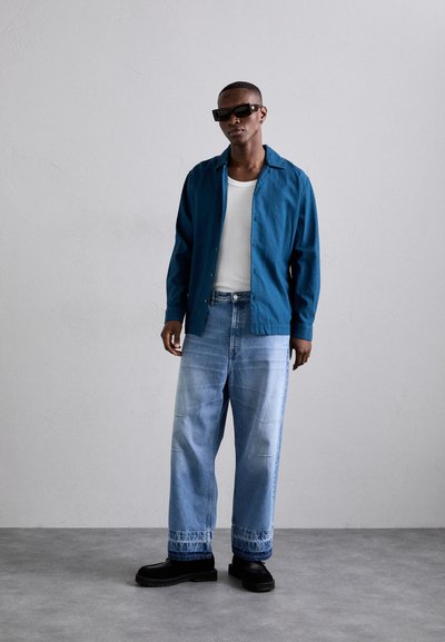 Chemise boutonnée turquoise, débardeur blanc, jeans larges en denim clair avec des poignets brodés et chaussures noires à semelles épaisses. Lunettes de soleil sombres portées.