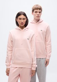 Ροζ φούτερ με κουκούλα από απαλό ύφασμα, με μπροστινή τσέπη καγκουρό και μικρό πράσινο λογότυπο. Το μοντέλο με γκρι παντελόνια στέκεται δίπλα σε άλλο με αντίστοιχα κάτω ρούχα.