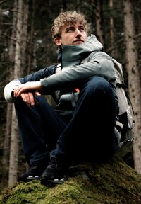 Athletic person sitting on a mossy rock, wearing a grey waterproof jacket, navy pants, and black hiking shoes, with a light backpack.