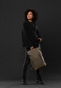 Olivgrüner Rolltop-Rucksack mit einer vorderen Reißverschlusstasche. Hergestellt aus strapazierfähigem Material. Das Modell trägt ein schwarzes Langarmshirt und enge Jeans.