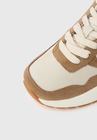 Αθλητικό παπούτσι σε μπεζ και καφέ χρώμα με ομαλή υφασμάτινη επάνω επιφάνεια, σουέντ λεπτομέρειες, στρογγυλή μύτη και ανοιχτόχρωμα κορδόνια. Καουτσούκ σόλα ορατή στο κάτω μέρος.