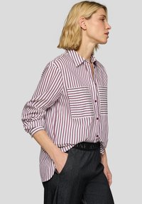Striped button-up shirt in white and maroon, featuring a collar, two front pockets, and a loose fit, paired with dark trousers.