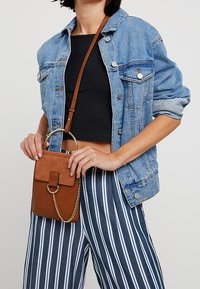 Ruskea nahkainen olkalaukku, jossa on pyöreä metallinen rengaskahva, varustettu läpällä ja ketjuakenteilla, yhdistettynä farkkutakkiin ja raidallisiin housuihin.