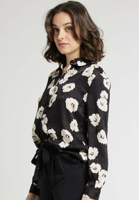 Blouse noire à fleurs avec des fleurs blanches et roses, manches longues, col et un lien à la taille. Tissu lisse avec un léger éclat.