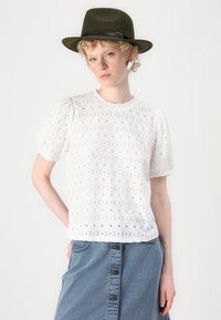 Witte kanten blouse met korte pofmouwen, een gestructureerd bloemenpatroon en een ronde halslijn, gecombineerd met een denimrok en een donkergroene hoed.
