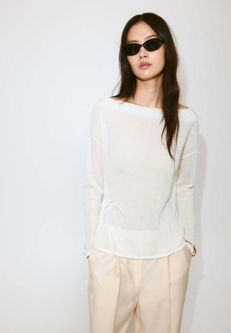 Femme portant des lunettes de soleil noires, un haut blanc transparent à manches longues et un pantalon beige clair, debout devant un fond blanc uni.