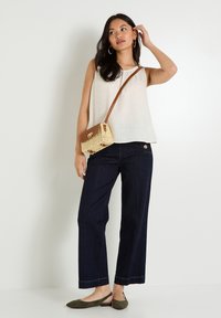 Haut crème léger sans manches avec une finition texturée ; jean large sombre ; chaussures plates vert olive ; sac bandoulière tissé avec des accents marron.