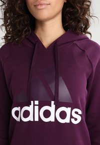 Sudadera con capucha púrpura que presenta un gran logotipo blanco de "adidas" y un acento triangular gris. Incluye cordones y un tejido suave y texturizado.