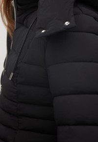 Zwarte gewatteerde jas met gestikte delen, een ritsluiting en drukknoopjes op de kraag. Glad textuur, geïsoleerd ontwerp en getailleerde pasvorm.