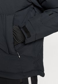 Schwarze gesteppte Jacke mit einer Reißverschlusstasche und Klettverschluss an den Manschetten. Der Stoff ist glatt mit einem gesteppte Muster und minimalen Metallteilen.