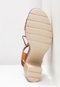 Semelle plate beige de sandale avec bande de roulement rainurée, brides en daim beige, petits clous en métal le long du bord et un talon bas en bois, incliné contre un mur blanc.