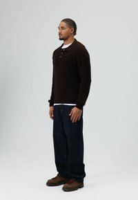 Man staat en draagt een donkerbruine gebreide trui, wit ondershirt, losse donkere spijkerbroek en bruine wandelschoenen tegen een eenvoudige lichte achtergrond.