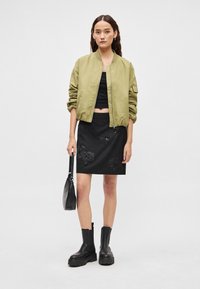 Olijfgroene bomberjack, zwart cropped topje, zwarte rok met bloemrijke borduursels, met een zwarte handtas, zwarte chunky enkellaarzen aan.