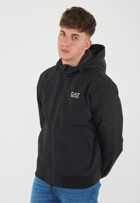 Black lightweight jacket with a front zipper, adjustable hood, and ribbed cuffs. Features a logo on the chest and streamlined design.
