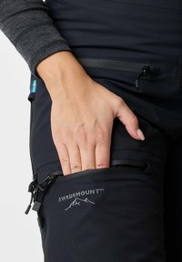 Mano che inserisce nella tasca con cerniera di pantaloni outdoor neri Swedemount con etichetta con bandiera svedese e camicia grigia a maniche lunghe visibile.