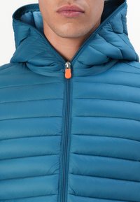 Jachetă căptușită în nuanță de teal, cu glugă, având închidere cu fermoar și un manson portocaliu. Model cu matlasare orizontală pe întreaga suprafață.