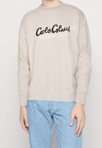 Beige Crewneck-Sweatshirt mit schwarzem Sticktext "Carlo Colucci" und "Milano 1978." Mit strukturierten Bündchen und Saum. Kombiniert mit hellblauen Jeans.