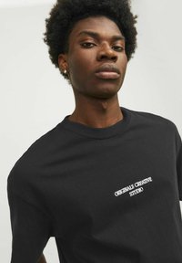 Zwarte katoenen t-shirt met korte mouwen, met daarop geborduurde witte tekst "ORIGINALS CREATIVE STUDIO" op de borst. Gladde textuur.