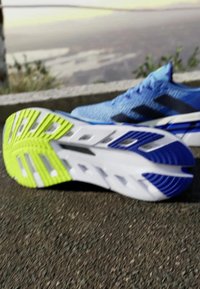 Blue athletic shoe with mesh upper, black stripes, and a textured grip sole. Notable details include a yellow-green outsole and white accents.