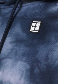 Tissu tie-dye bleu avec cordon noir, comportant un petit patch rectangulaire avec une bordure noire et un symbole abstrait en noir et blanc.