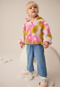 Veste en polaire avec des motifs de taches roses et jaunes, fermeture éclair, associée à un jean large bleu et des baskets multicolores.