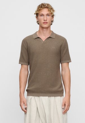Jonge man met krullend blond haar, gekleed in een bruin gebreid poloshirt met korte mouwen en lichtbeige geplooide broek, staand tegen een effen achtergrond.