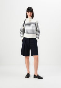 Donna che indossa un maglione a righe bianche e blu scuro con bottoni, pantaloni corti neri fino al ginocchio, mocassini neri e porta una borsa a tracolla con motivo.