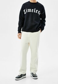 Hombre vestido con un suéter negro con el texto "Timeless", pantalones blanco roto y zapatillas en blanco y negro, de pie frente a un fondo blanco liso.